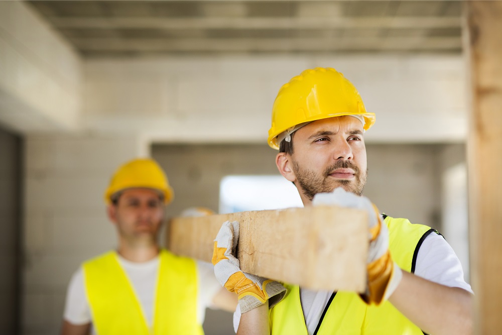 Two construction workers wearing safety helmets and gloves carrying a wooden beam. 